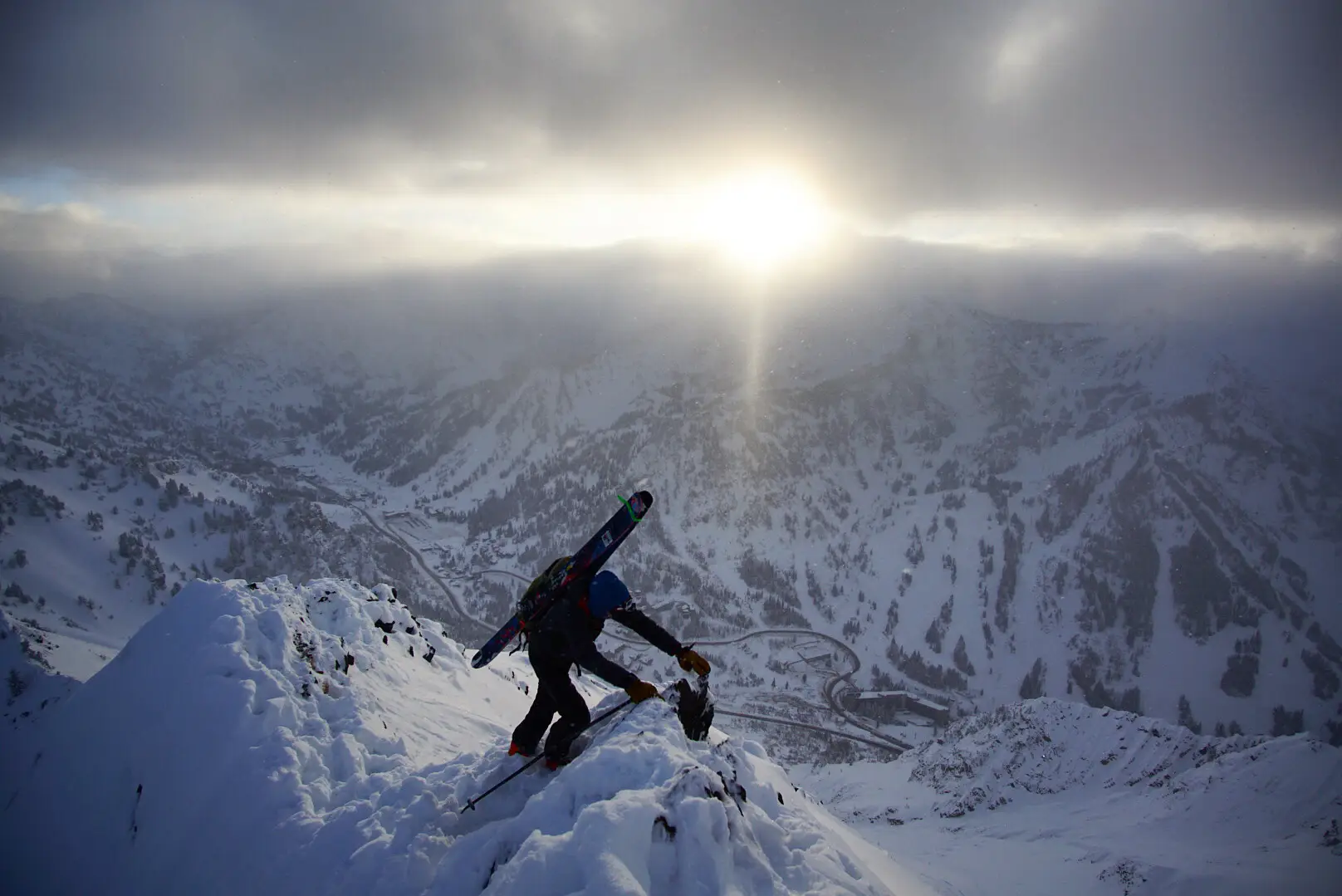 Skier hiking up a snowy mountain ridge with skis on their back at sunrise.