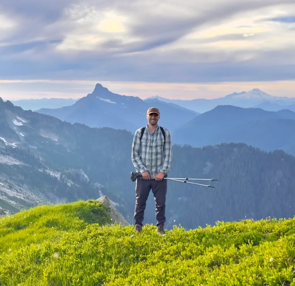 Person standing on a grassy mountain ridge holding trekking poles with layered mountain peaks in the distance.