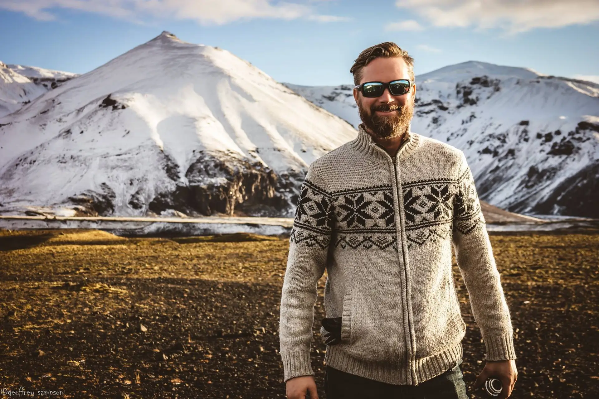 Man wearing a patterned sweater and sunglasses standing in front of snow-covered mountains.