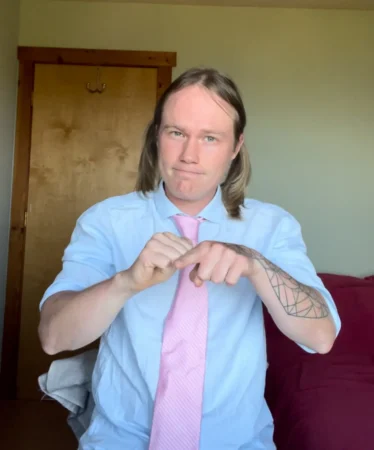 Person wearing a light blue shirt and pink tie making a hand gesture while standing indoors.