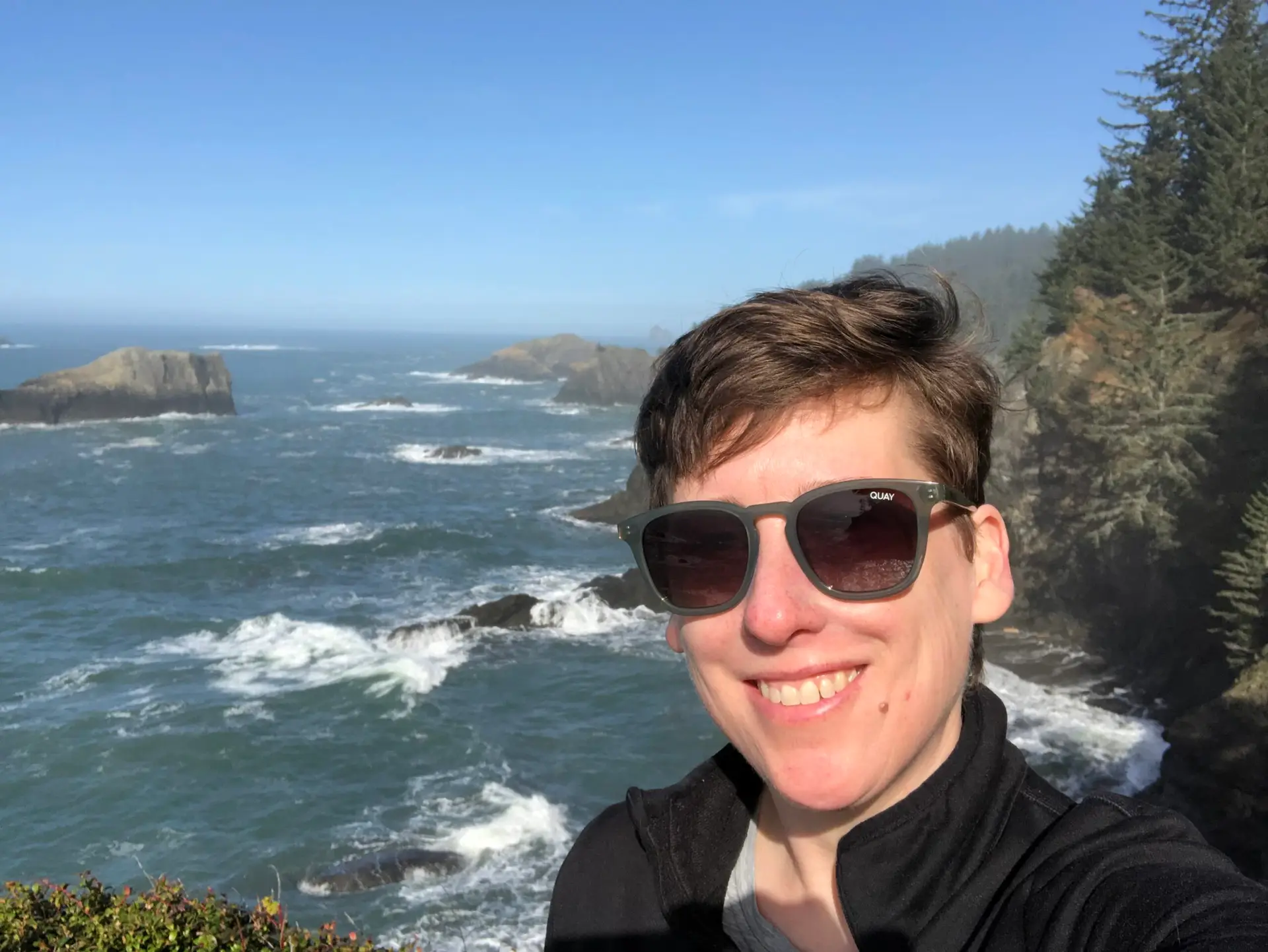 Woman wearing sunglasses smiling in a selfie with ocean waves and rocky coastline behind her.