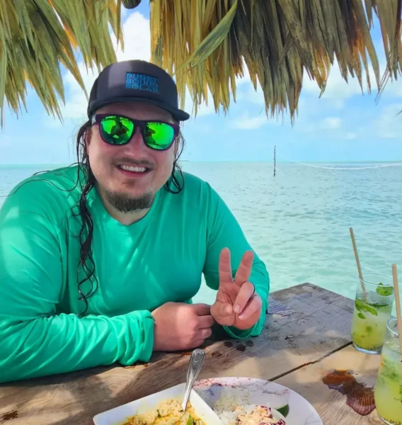 Person wearing a green shirt and sunglasses makes a peace sign while seated at a beachside table with food and drinks.
