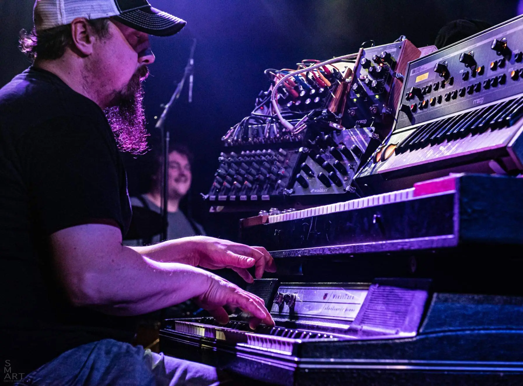 Musician wearing a cap playing a keyboard and synthesizer setup under purple stage lighting.