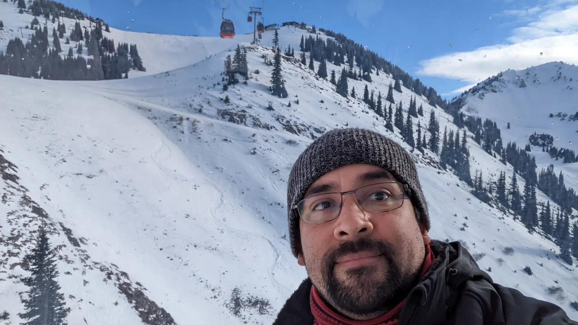 Person wearing a knit beanie and glasses with snowy mountain slopes and gondola cars in the background.