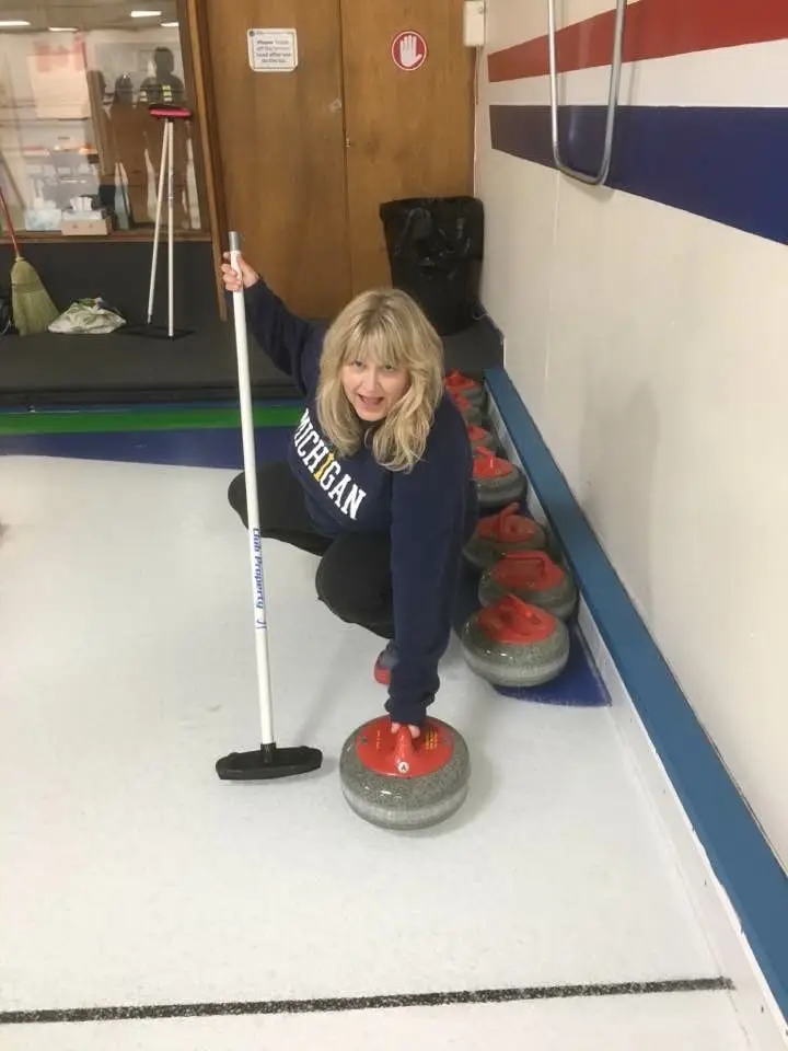 Woman crouching on an indoor curling rink holding a broom and a curling stone.