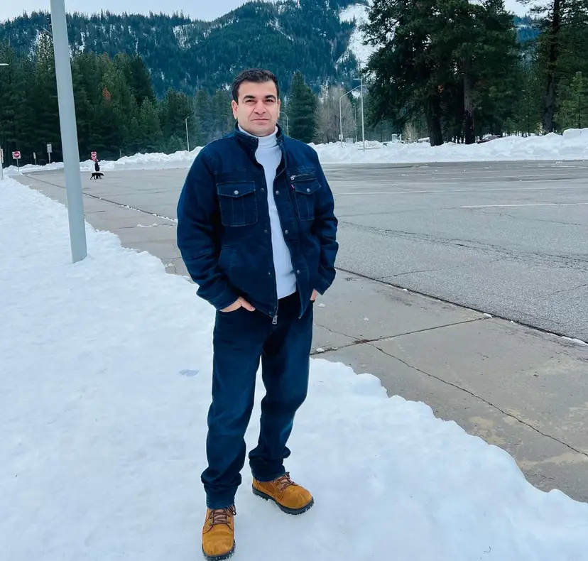 Person standing on a snowy roadside wearing a denim jacket, jeans, and tan boots with mountains and trees in the background.