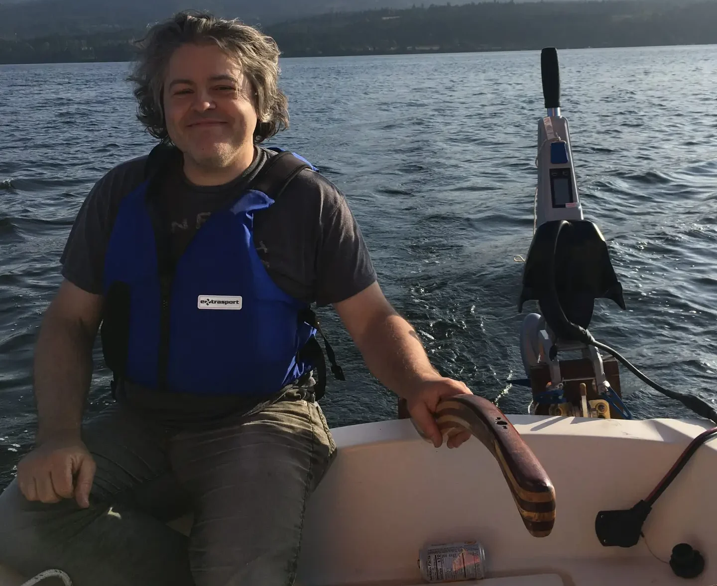 Smiling person sitting in a small boat on a lake with mountains in the background.