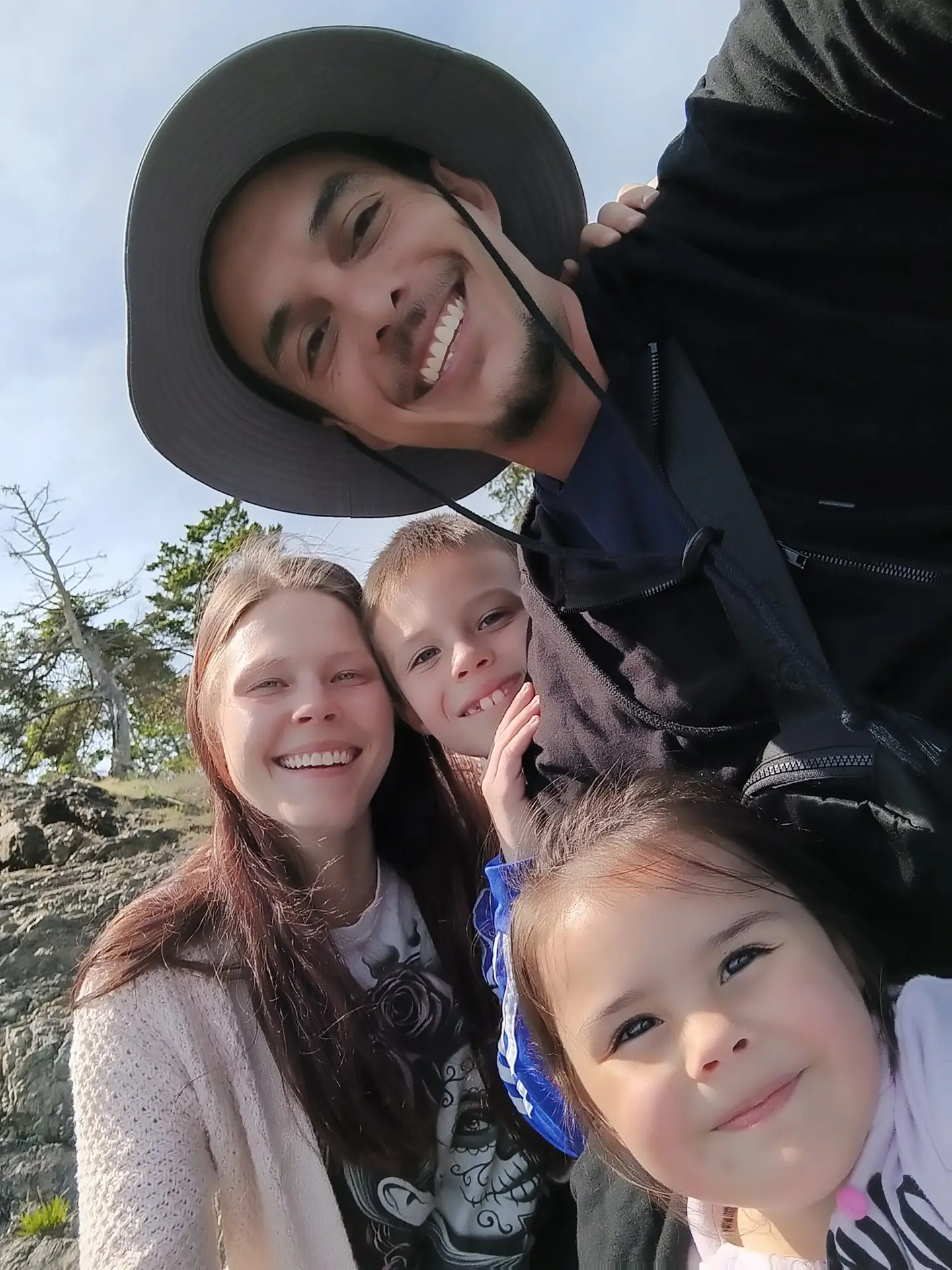 Smiling family of four taking a selfie outdoors on a sunny day.