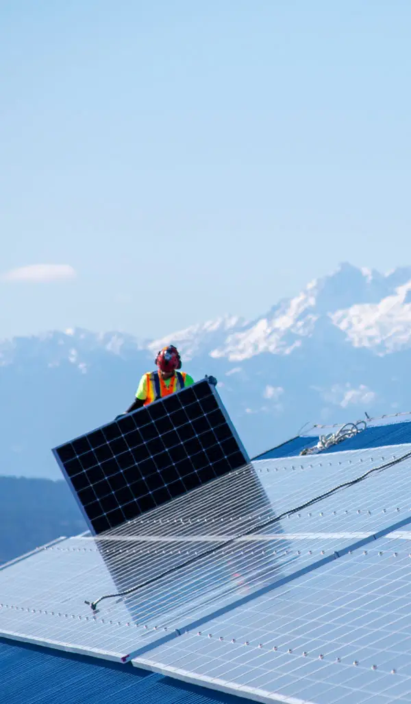Solar installer lifting a photovoltaic panel onto a rooftop with mountains in the background.