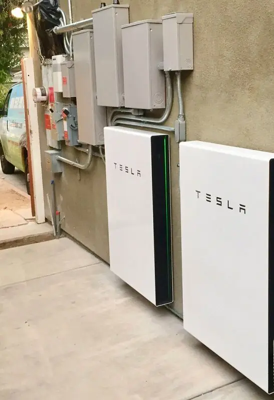 Two Tesla Powerwall batteries mounted on an exterior wall with electrical panels.