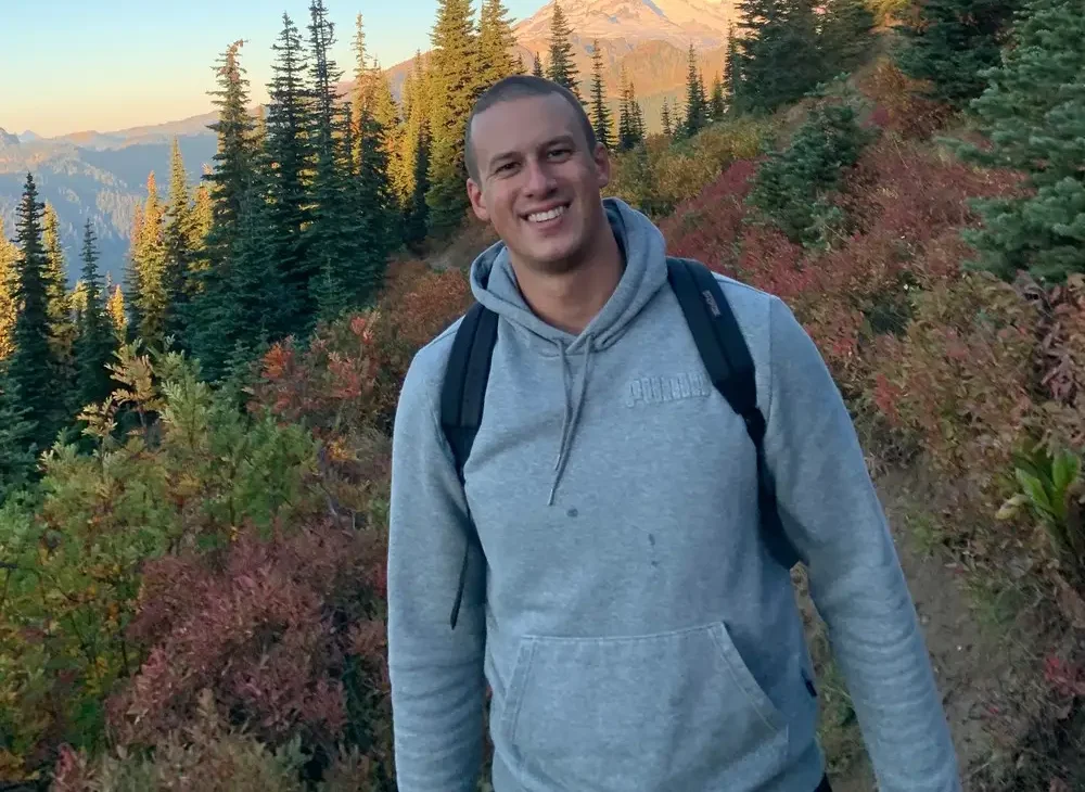 Smiling man in a gray hoodie and backpack standing on a mountain trail surrounded by trees